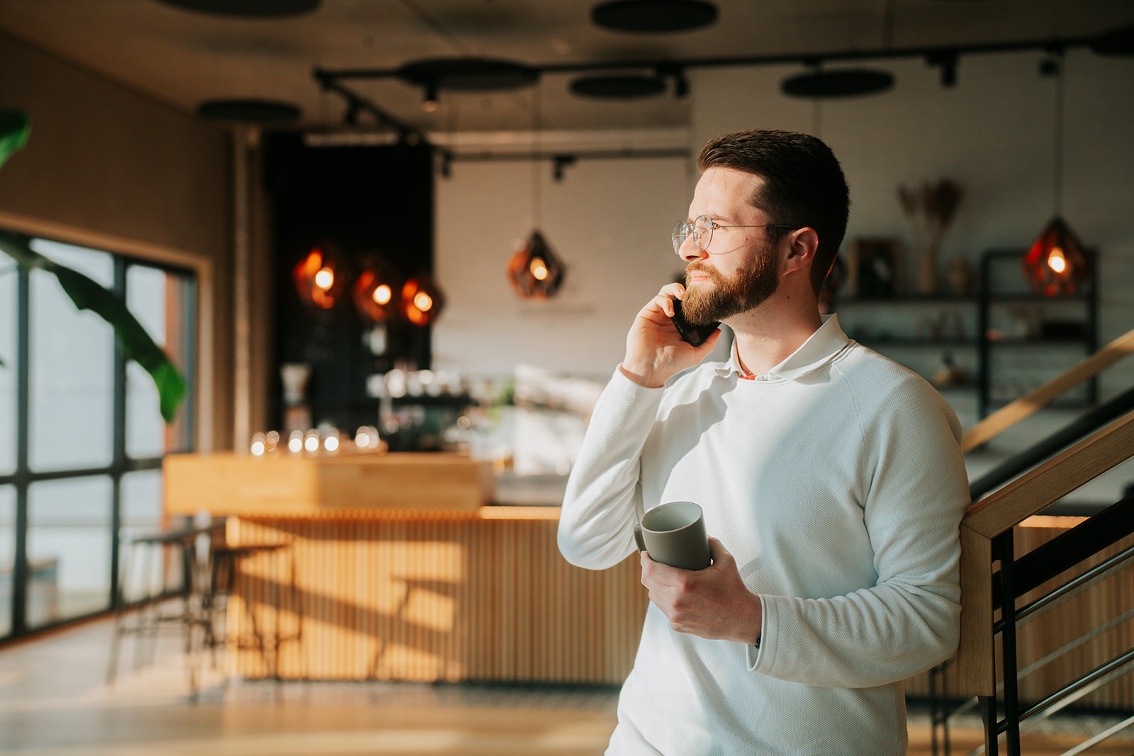 Ein junger Mann mit Bart und Brille telefoniert in einem modernen Café oder Co-Working-Space, während er eine Kaffeetasse hält.
